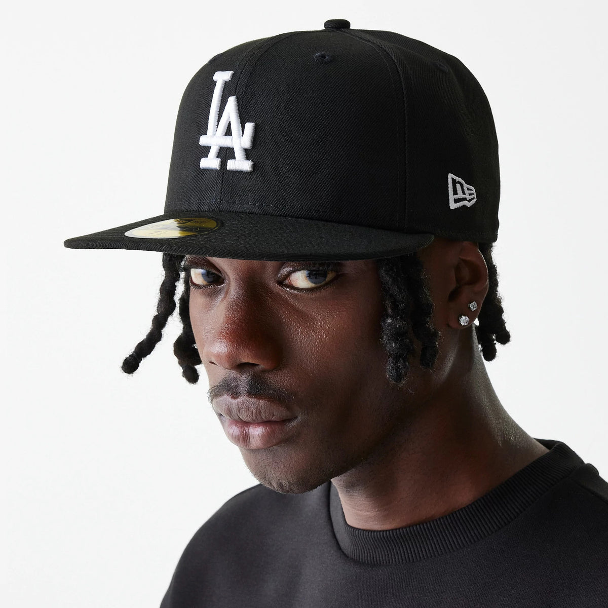 LA Dodgers Essential Black New Era 59FIFTY Cap, on-model view with a white raised embroidered LA logo on the front panel, gold 59FIFTY size sticker on the flat visor, and the New Era flag logo on the left side above the ear.