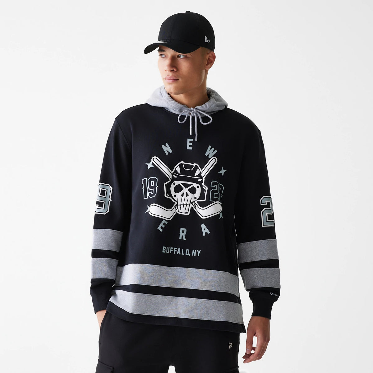 New Era Street Dangle Grey Pullover Hoodie, front view showing a large skull and hockey theme printed centrally, black sleeves with grey stripes, and a grey drawstring hood, worn by a person also in a black cap with an embroidered New Era logo.