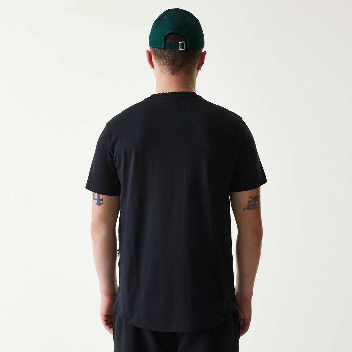 New Era Outdoor Earth Day Black T-Shirt, on-model rear view showing the plain black fabric extending over the shoulders, hem finishing below the waist, and a green cap with a debossed New Era logo on the back of the head.