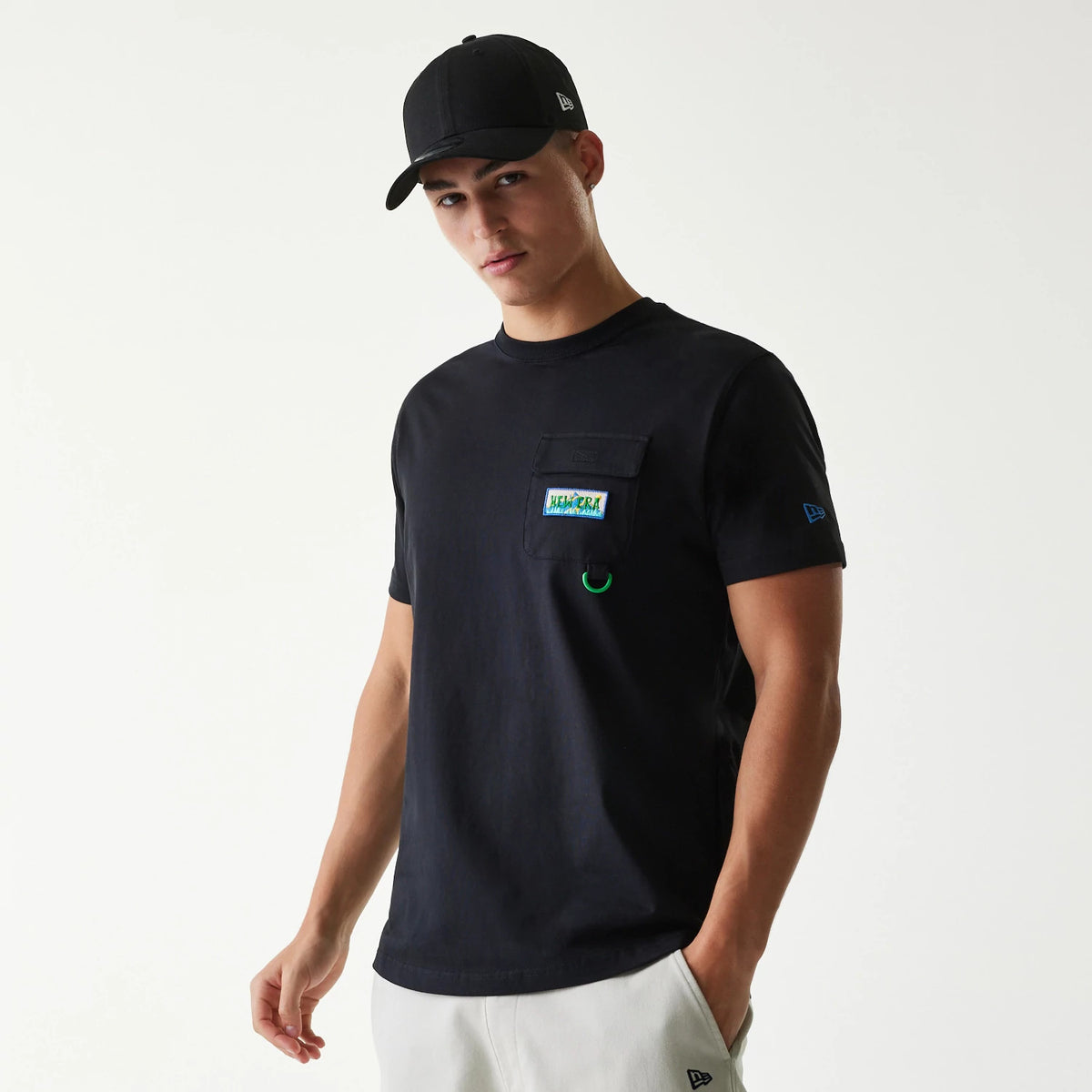 New Era Outdoor Earth Day Black Side Pocket T-Shirt, on-model view showing the black tee with a debossed square pocket on the chest, a colourful Earth Day patch above the pocket, and a blue New Era logo on the left sleeve.