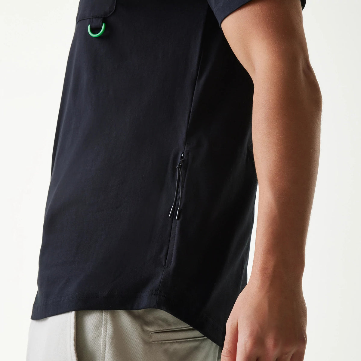 New Era Outdoor Earth Day Black Side Pocket T-Shirt, side view showing a sleek black design with a zip pocket, short sleeves ending just above the elbow, a small green loop near the shoulder and the hem sitting above light-coloured trousers.