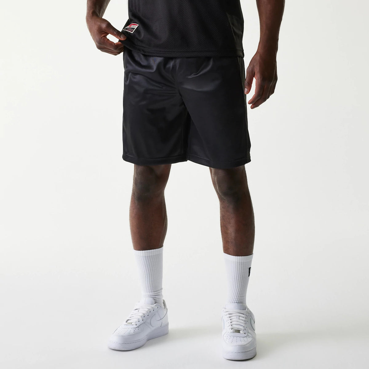 New Era Retro Sports Black Mesh Shorts, front view showing the plain black shorts worn with a waist label exposed, ending above the knee, and paired with tall white socks and white trainers.