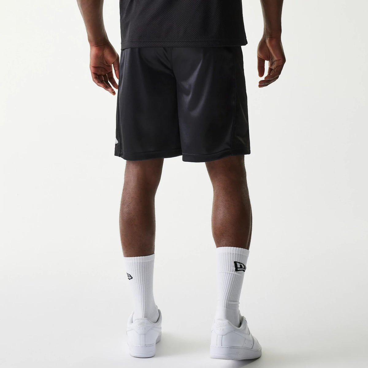 New Era Retro Sports Black Mesh Shorts, rear view showing the loose fit black shorts with a debossed New Era logo on the lower right leg near the hem, worn by a model also wearing white socks and sneakers.