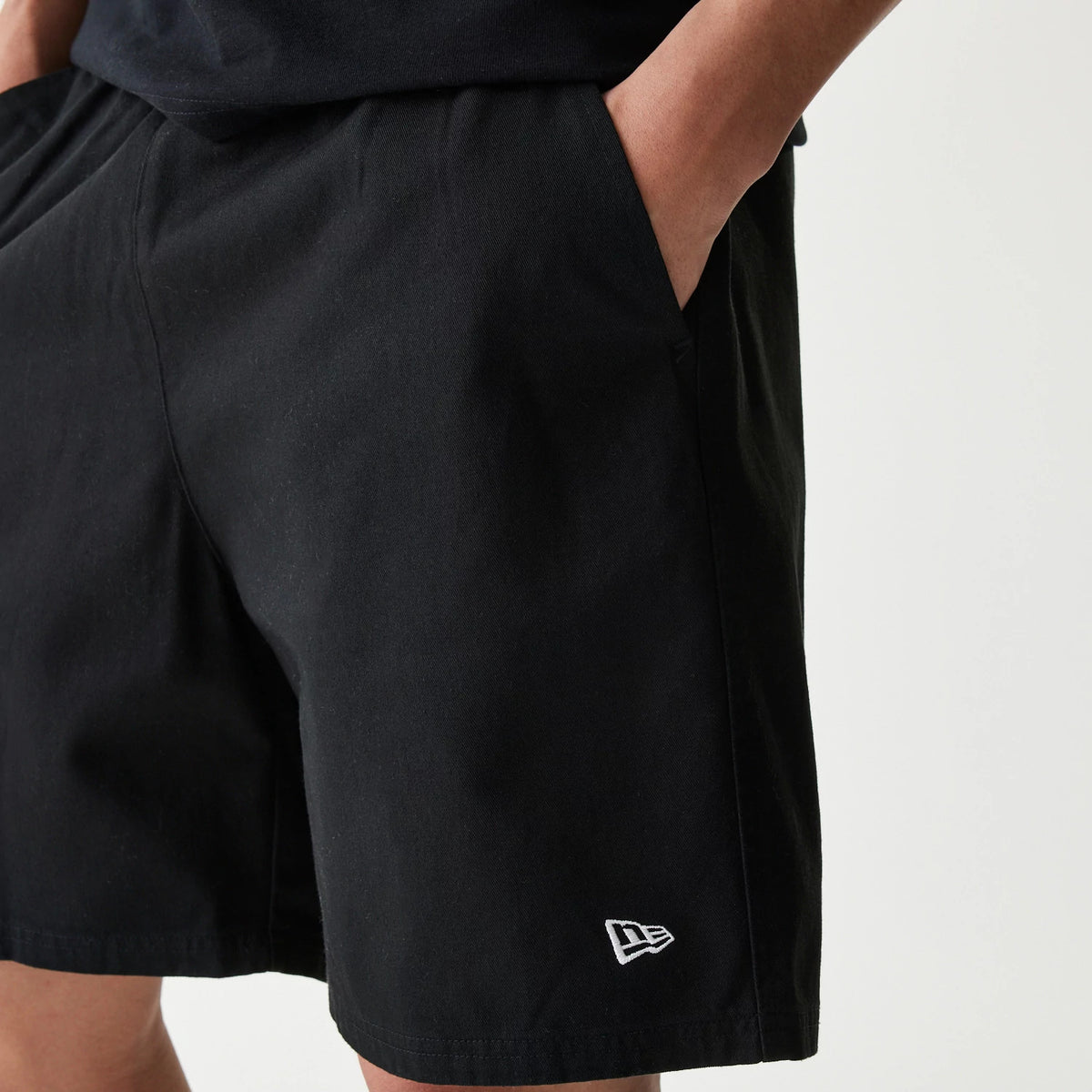 New Era Japan Woven Black Shorts, front view showing the plain black fabric with a white embroidered New Era logo on the lower left leg above the hem, hands casually placed in side pockets.