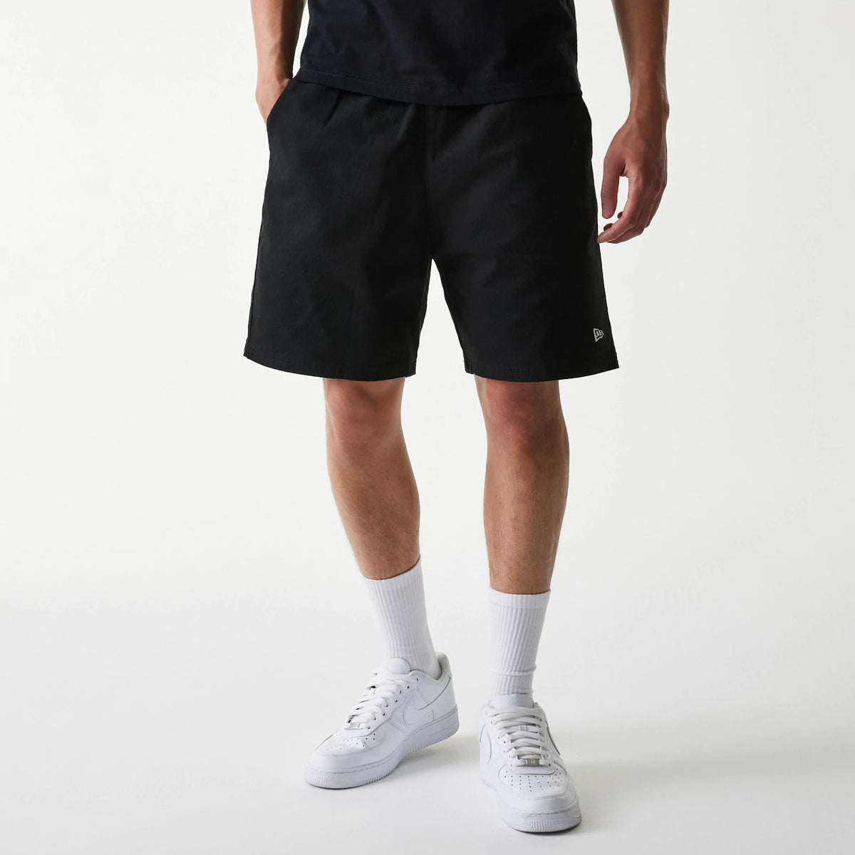 New Era Japan Woven Black Shorts, front view showing the plain black fabric with a small embroidered New Era logo in white near the hem on the left leg, styled with white trainers and socks.