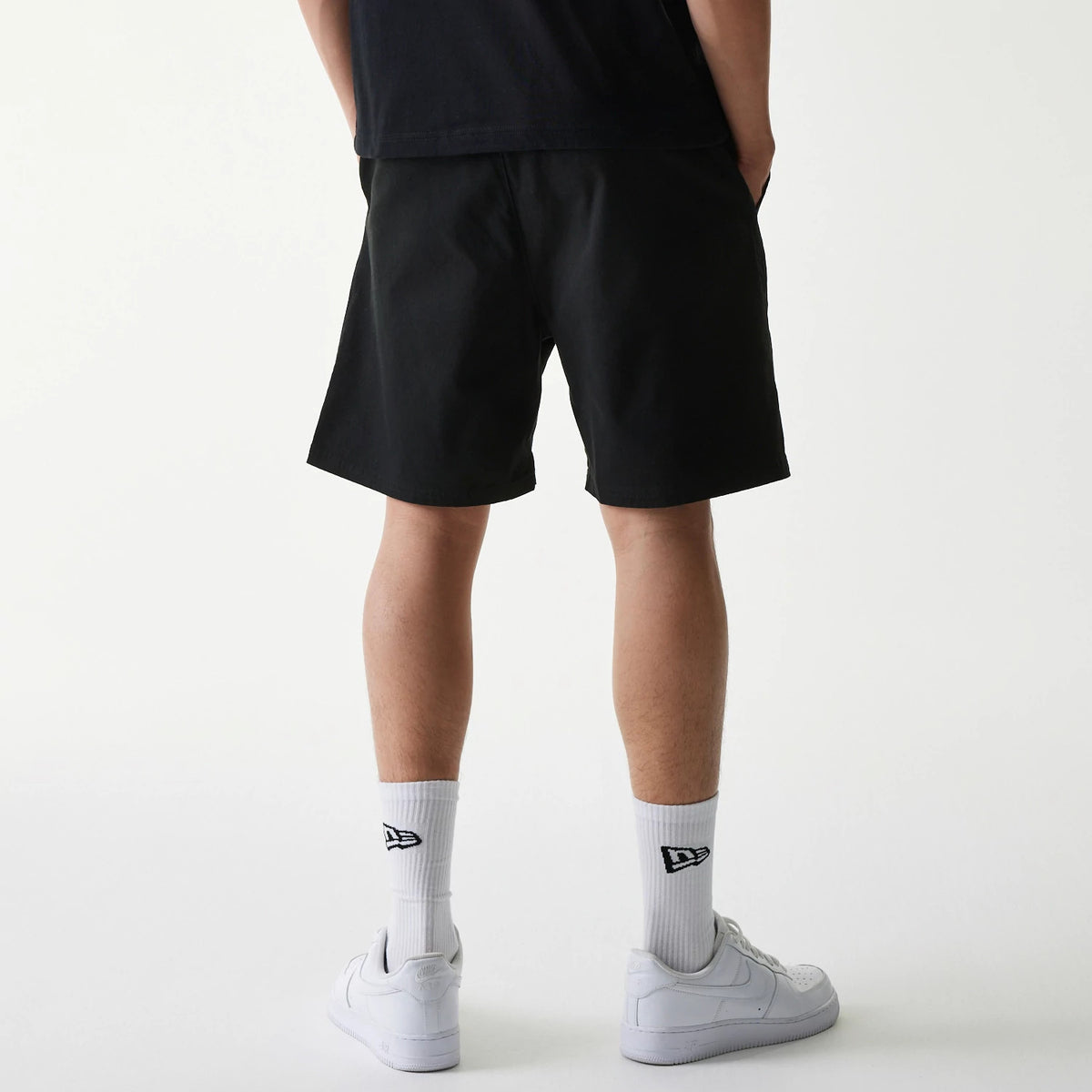 New Era Japan Woven Black Shorts, rear view displaying the plain black fabric on-model, with a simple design reaching just above the knee, paired with white socks featuring an embroidered New Era logo above white trainers.