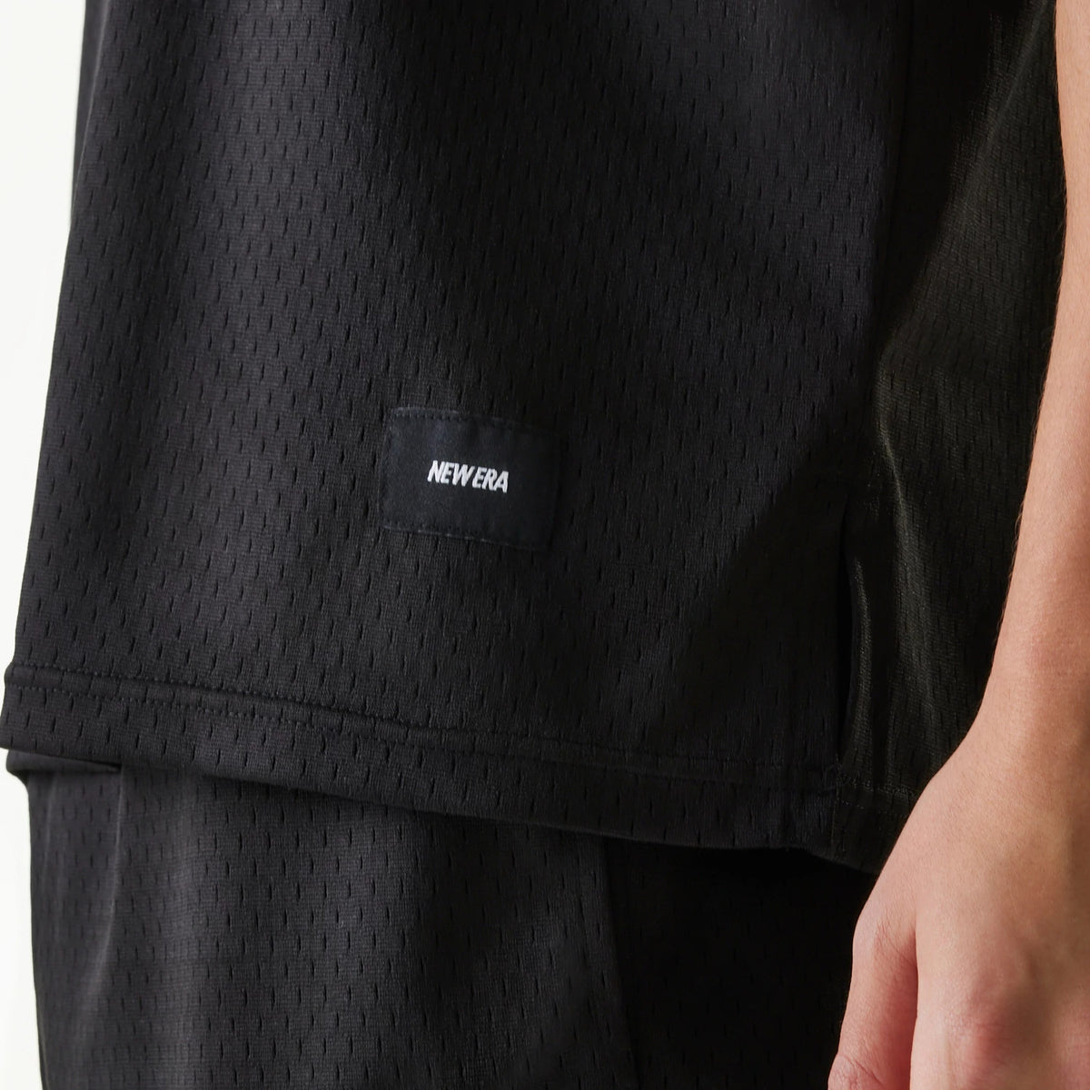 New Era Japan Mesh Black Sleeveless T-Shirt, side view showing the black mesh fabric with a raised New Era logo tag on the lower left side above the hem, seamless arm opening, and a close-up of the right side hip.