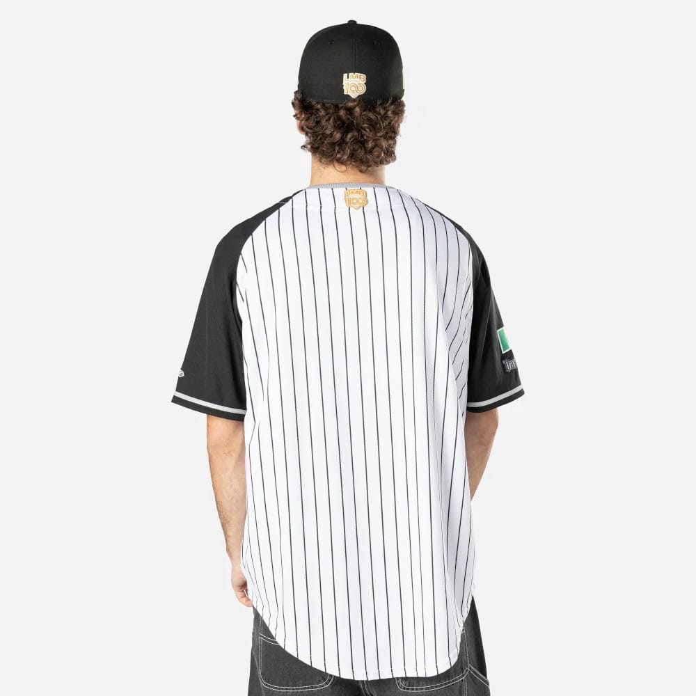 Oaxaca Guerreros Mexico LMB On Field White On-Field Jersey, back view on a model with a black cap, featuring vertical pinstripes and a golden LMB 100 logo on the upper back, complemented by black sleeves with white trim.