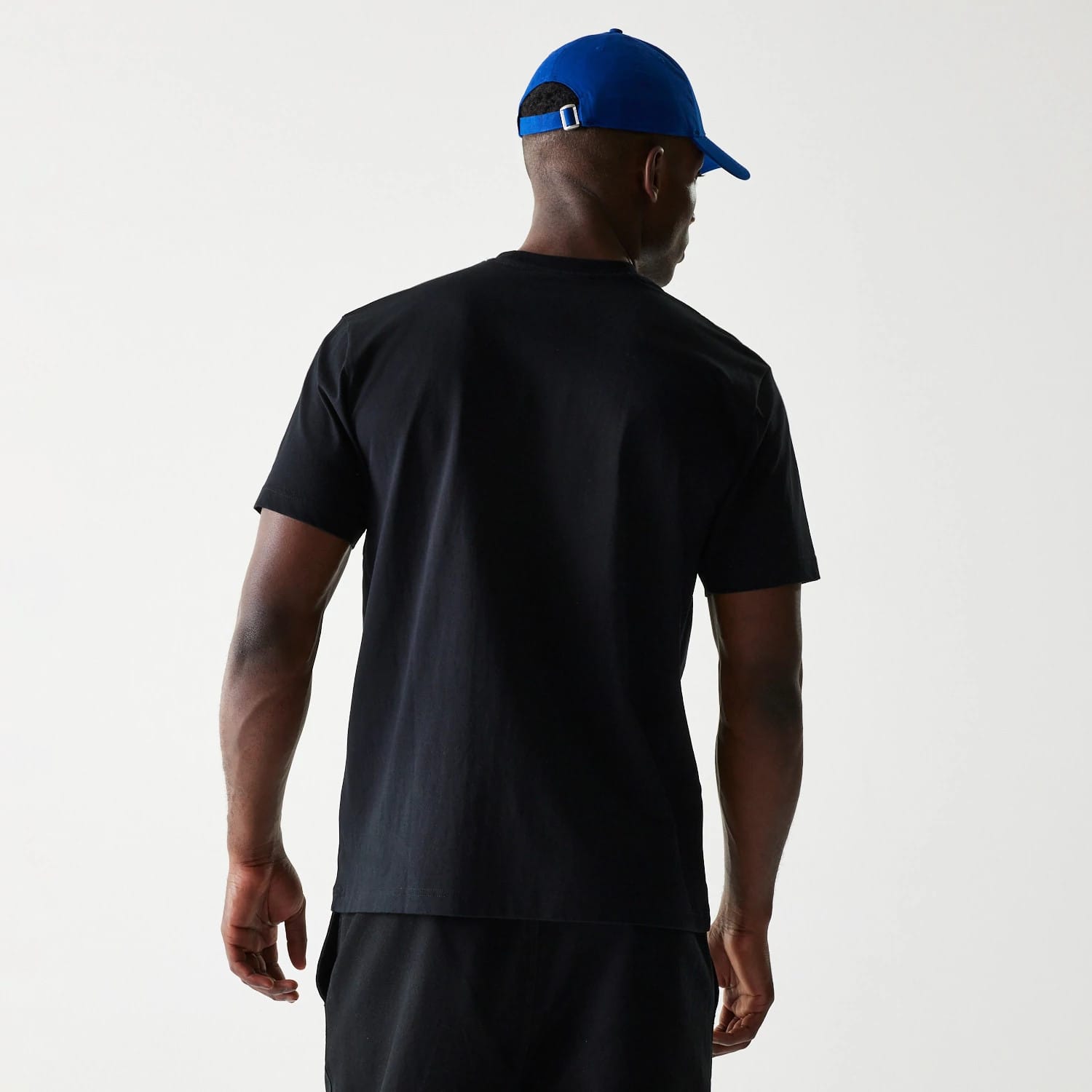 LA Dodgers MLB Paisley Black Oversized T-Shirt, rear view showing the plain black back with short sleeves and a relaxed fit, worn by a person in a blue cap with a debossed New Era logo on a purple cap.