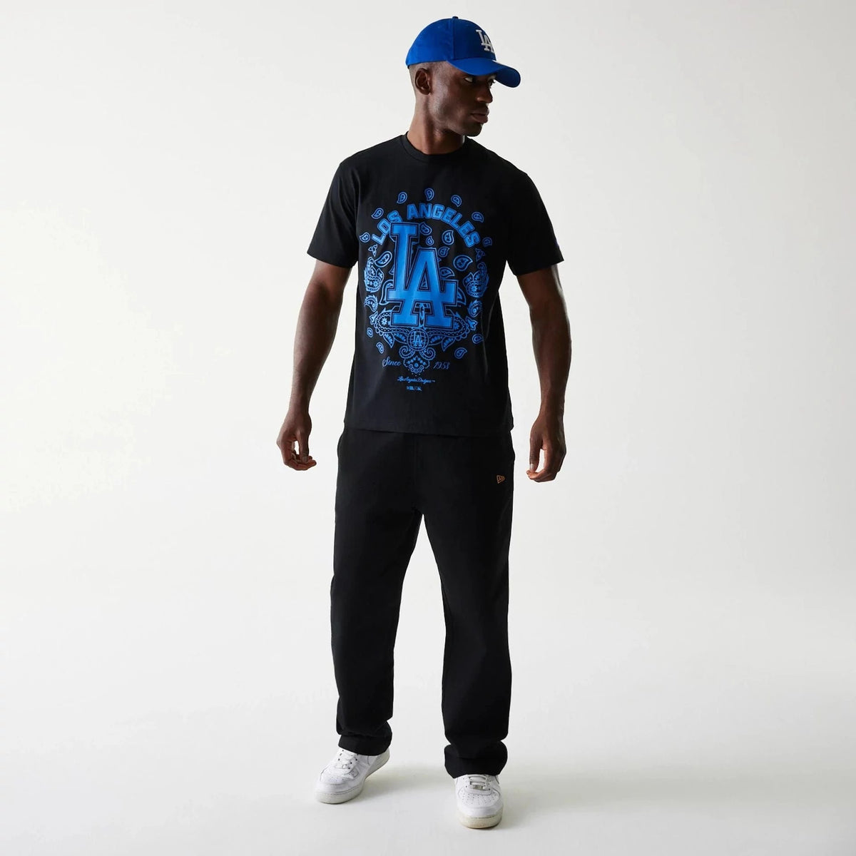 LA Dodgers MLB Paisley Black Oversized T-Shirt, on-model view showing a man wearing a black T-shirt with a large blue LA logo and intricate paisley design on the chest, paired with black trousers and a blue cap with a white embroidered LA logo.