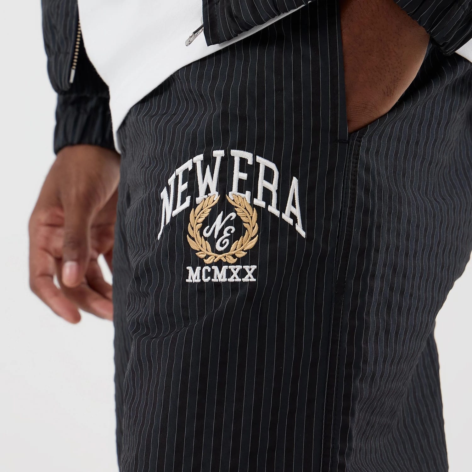 New Era Street Class Navy Straight Leg Joggers, close-up with a pinstripe design featuring white and gold embroidered NEW ERA graphics and MCMXX lettering on the upper leg, styled with a white top and matching jacket.