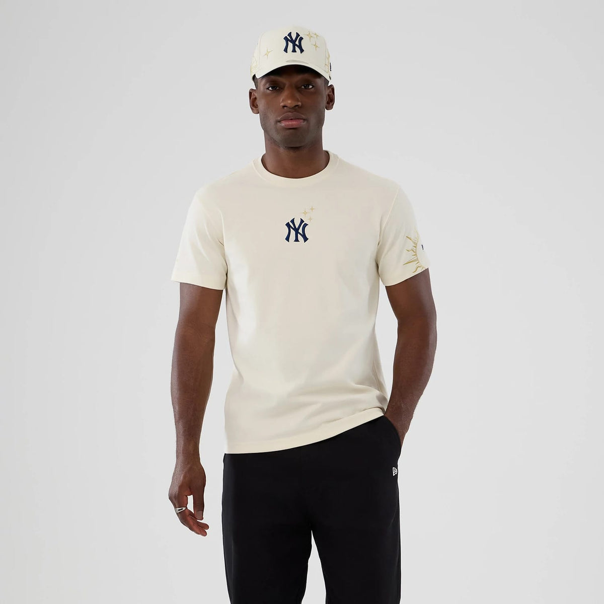New York Yankees MLB Cosmic Craft Cream T-Shirt, on-model view showing a cream tee with a navy New York Yankees logo centred on the chest, gold star accents around the emblem, and matching cap worn by a model.