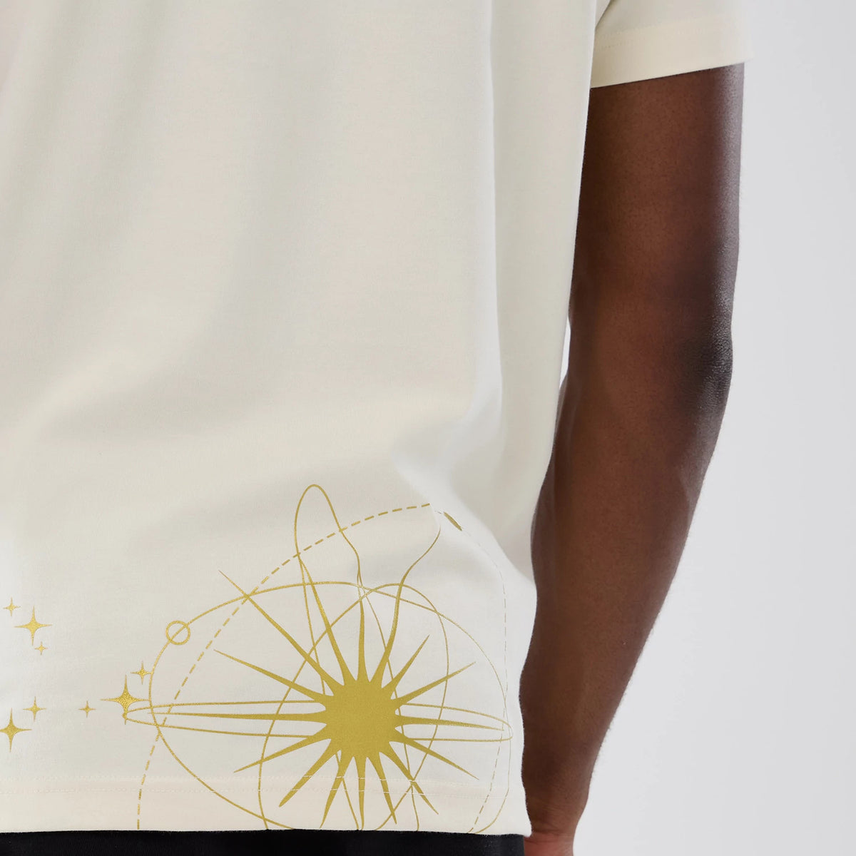 New York Yankees MLB Cosmic Craft Cream T-Shirt, side view showing the cream fabric with a golden cosmic design near the lower back hem, short sleeves, and the edge of the arm of the person wearing it.