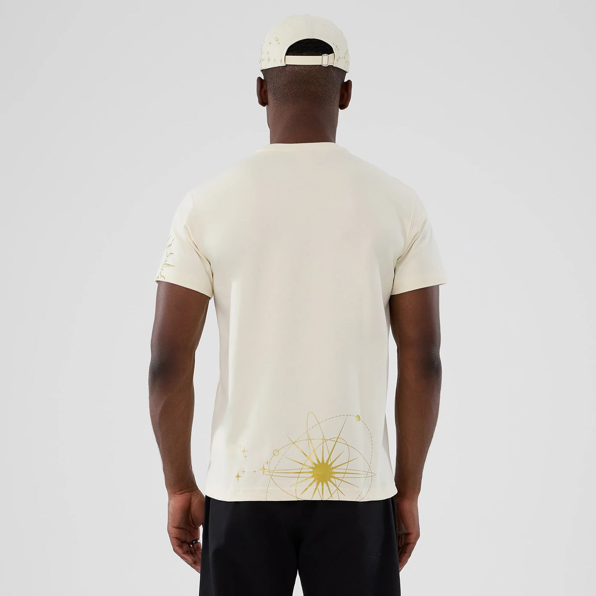 New York Yankees MLB Cosmic Craft Cream T-Shirt, rear view showing the cream fabric with a gold atomic design near the hem, short sleeves, and a cream cap with a debossed logo on a purple cap.