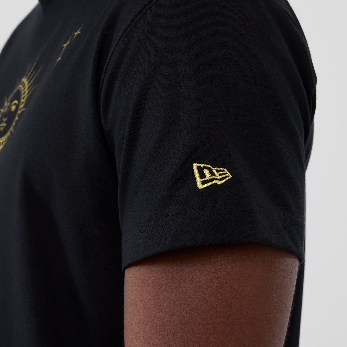 New Era Cosmic Craft Black T-Shirt, side view showing a close-up of the sleeve with a small raised yellow New Era logo embroidered near the hem and part of a celestial design in yellow on the black fabric.