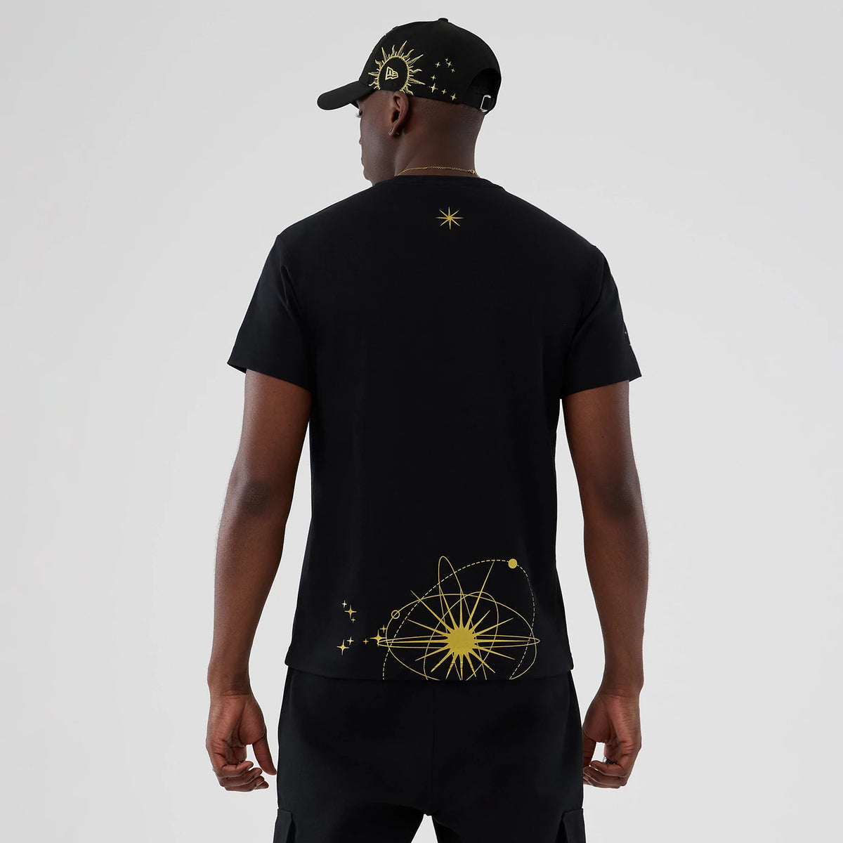 New Era Cosmic Craft Black T-Shirt, rear view showing a black shirt with a large yellow cosmic design at the lower back and a smaller star near the neckline, worn by a model in a black cap with a similar design.