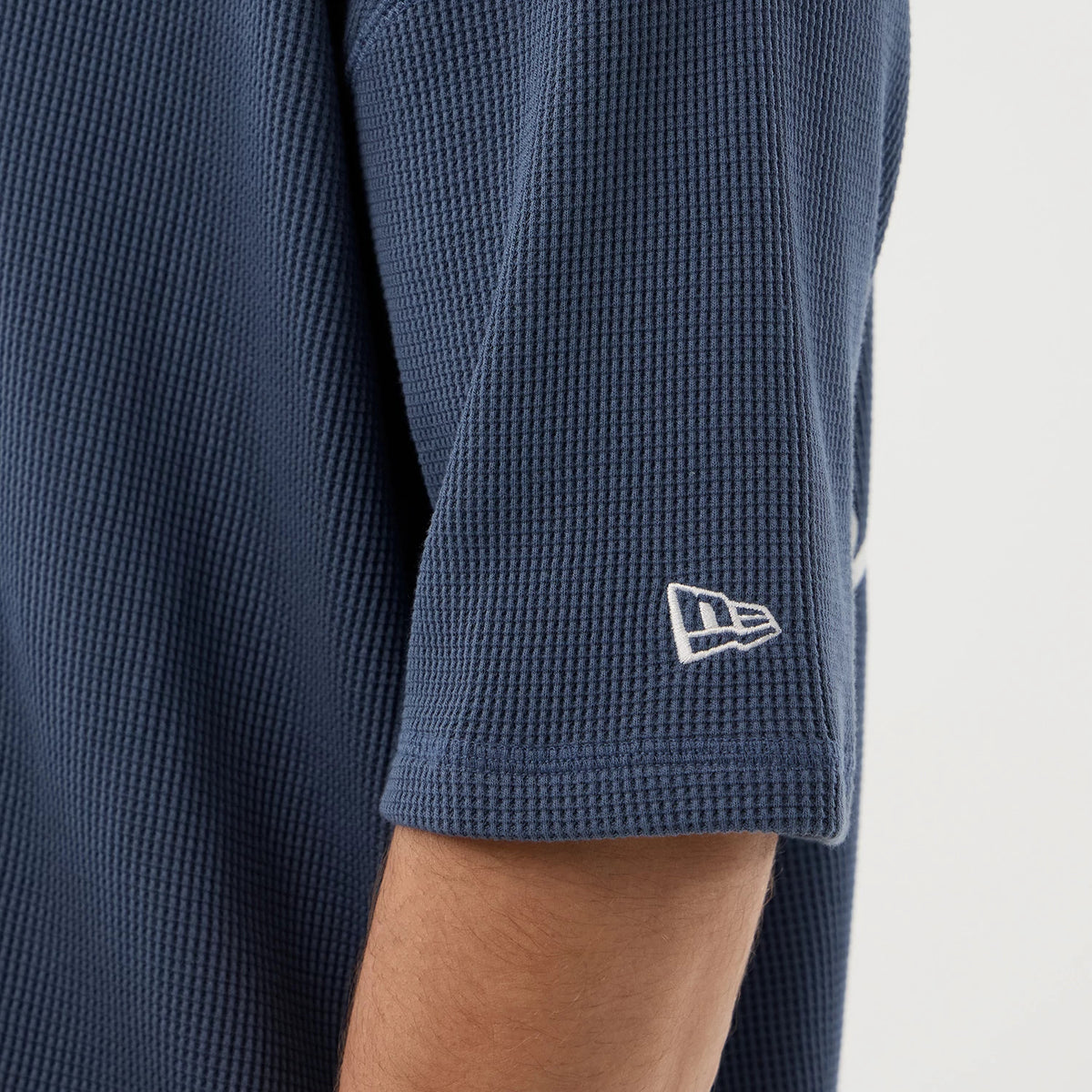 New Era Basic Blue T-Shirt, side view showing the textured blue fabric of the sleeve with a white embroidered New Era logo near the hem, displaying the clean edge and minimal design of the garment.