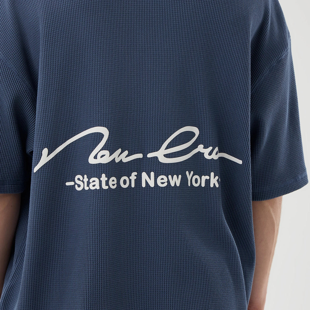 New Era Basic Blue T-Shirt, rear view showing the white printed script logo with “State of New York” text across the upper back on a textured blue fabric, short sleeves extending to the sides.