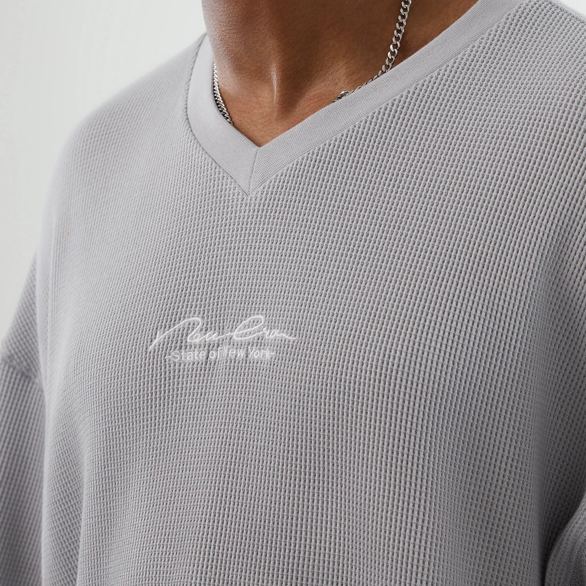 New Era Basic Grey T-Shirt, front view showing the light grey fabric with a V-neckline and an embossed signature logo in white on the chest, featuring a waffle knit texture around the shoulders and upper chest.