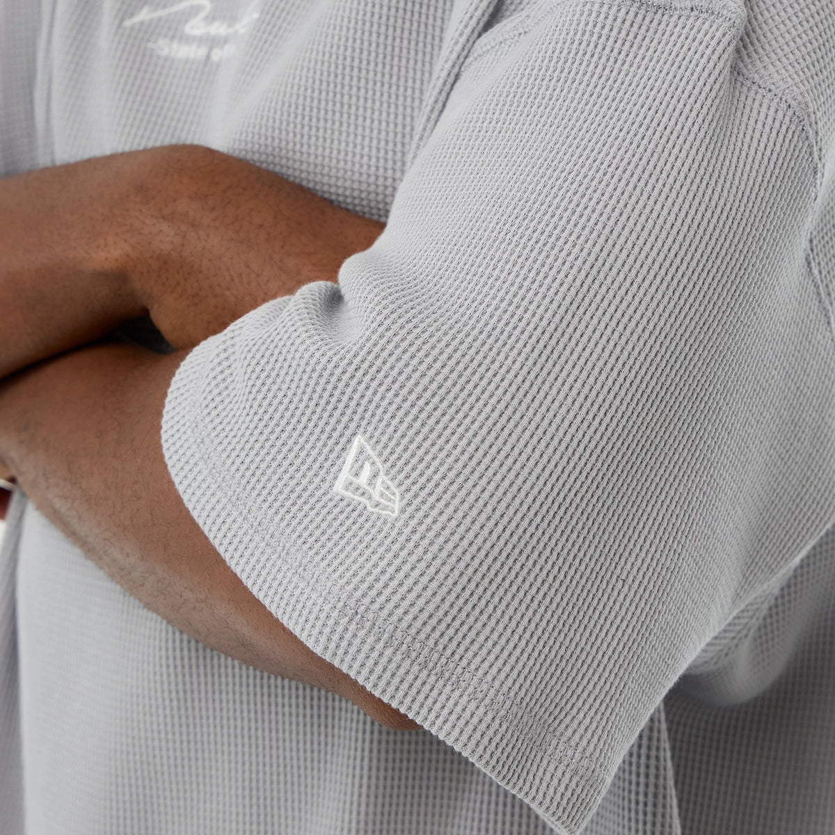New Era Basic Grey T-Shirt, side view showing the textured grey short sleeve with a small white embroidered New Era logo near the hem and the edge of the sleeve curving around the arm.