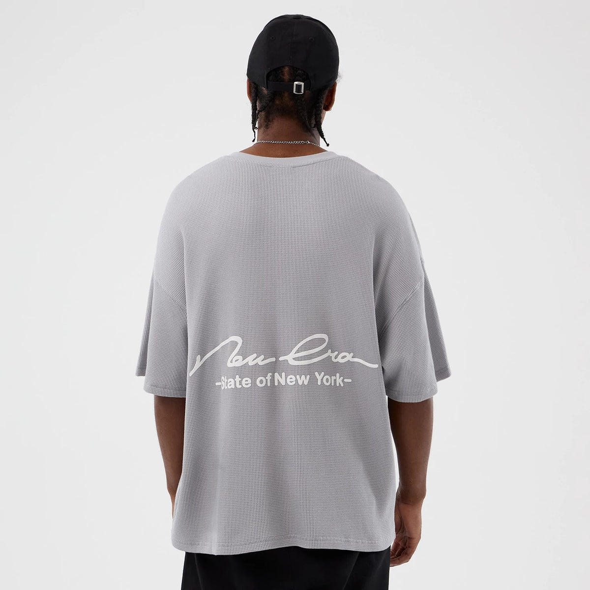 New Era Basic Grey T-Shirt, rear view showing the oversized grey fit with a white embossed New Era logo and "State of New York" text across the back, worn by a person also wearing a black cap.