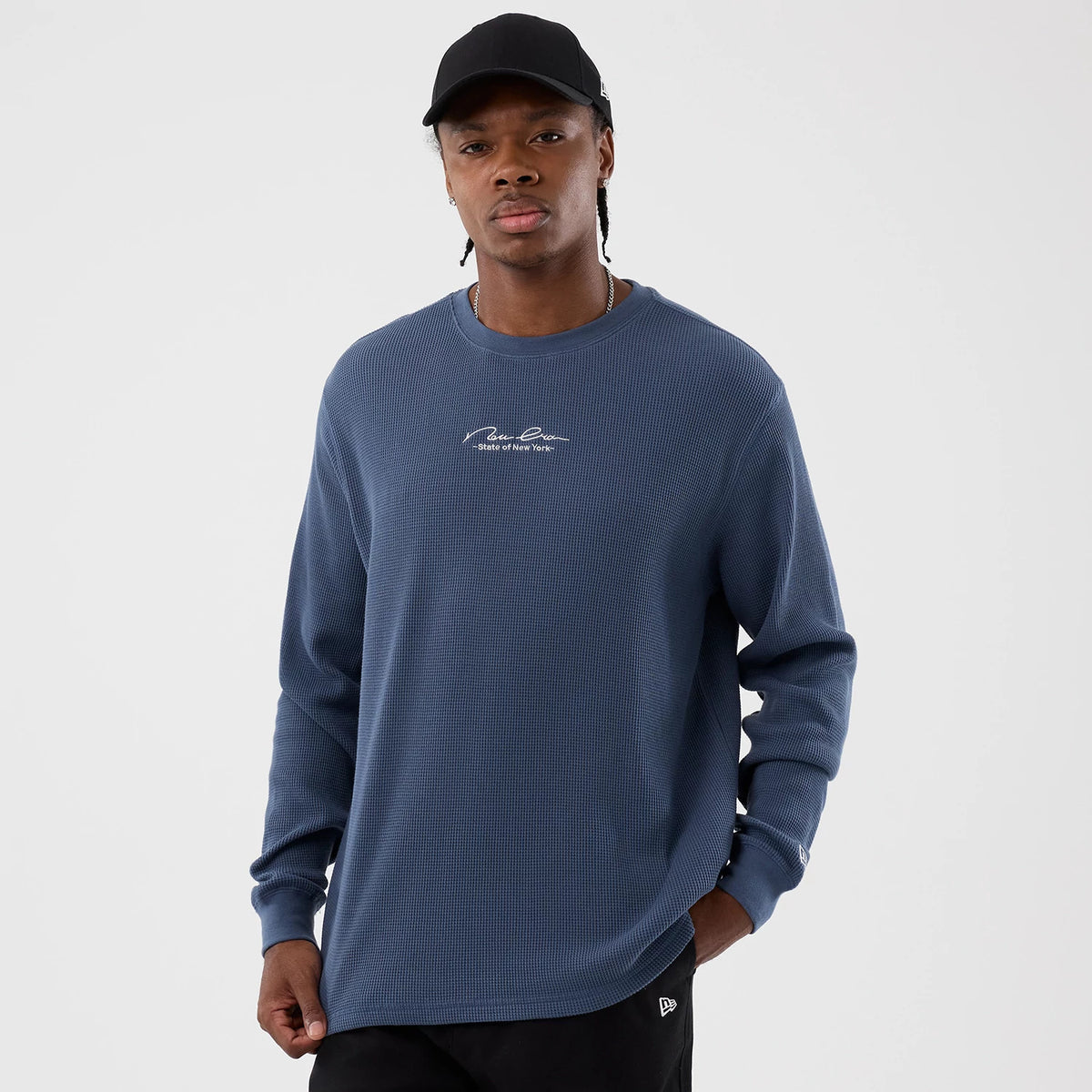 New Era Basic Blue Long Sleeve T-Shirt, front view showing a relaxed fit in blue with white script across the chest and a debossed logo on the left sleeve, worn by a model with hands in pockets.