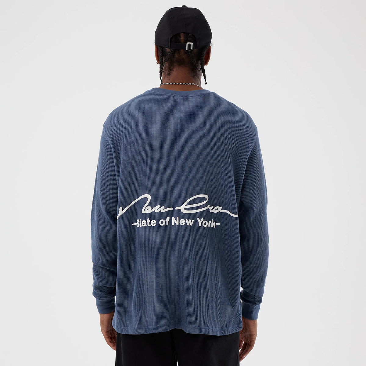 New Era Basic Blue Long Sleeve T-Shirt, rear view showing the blue fabric with a large white embossed New Era logo and "State of New York" text across the upper back, worn by a person with a black cap.