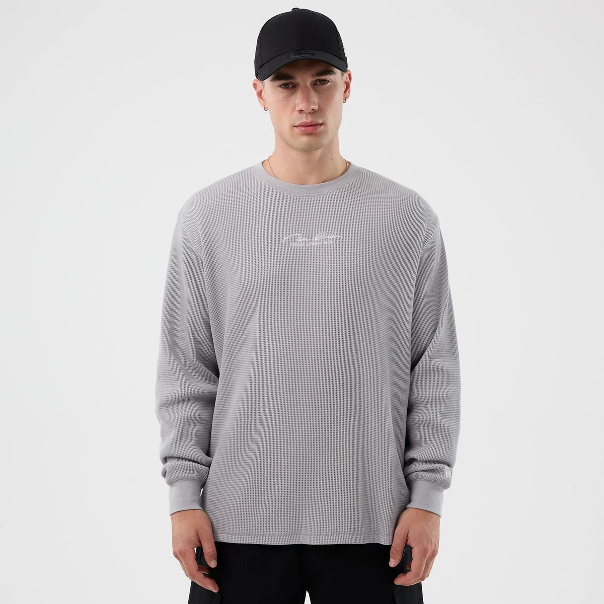 New Era Basic Grey Long Sleeve T-Shirt, front view showing a relaxed fit grey crew-neck with long sleeves, minimal text logo centred on the chest, worn by a model wearing a black cap.