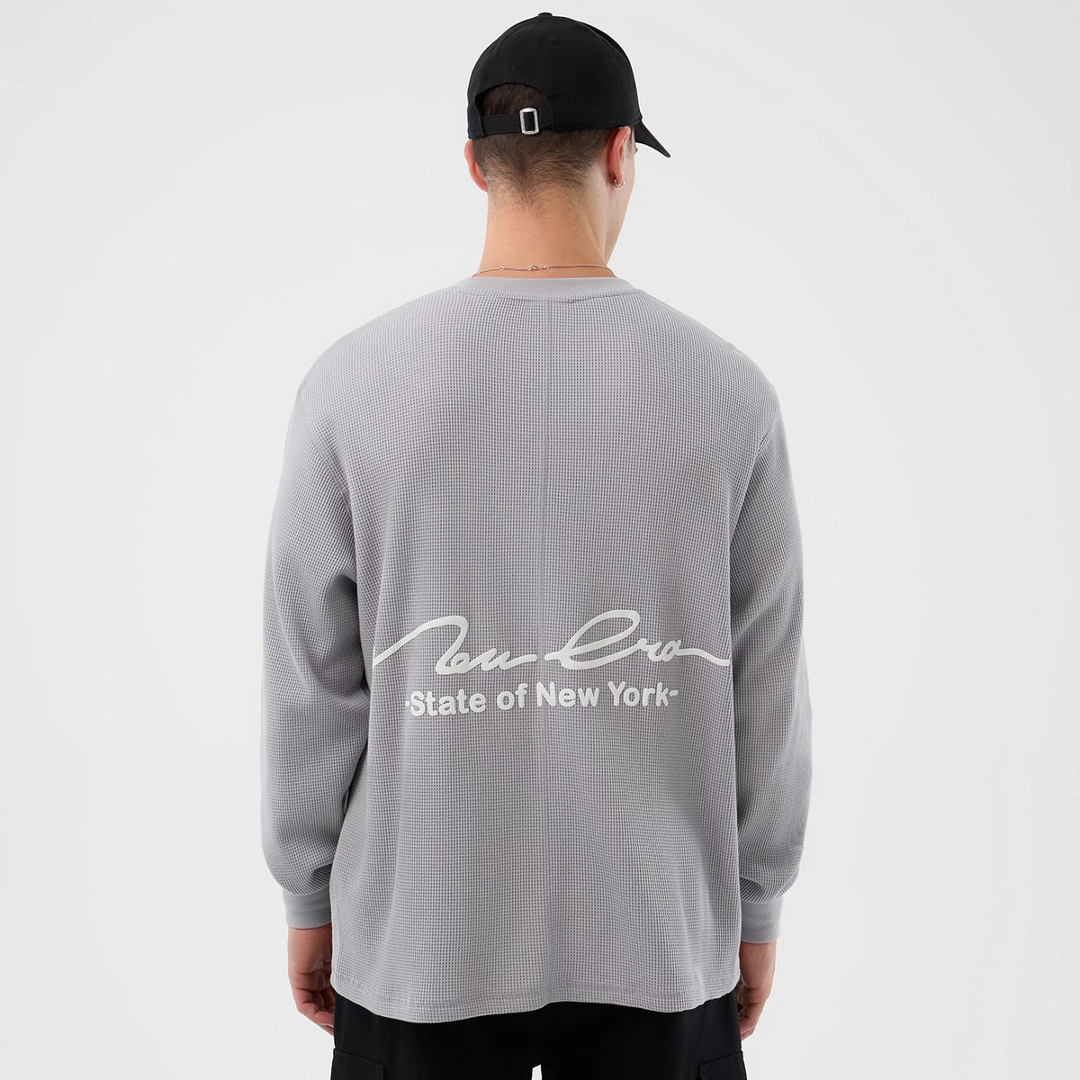 New Era Basic Grey Long Sleeve T-Shirt, rear view showing the grey textured fabric with a large white scripted New Era logo and "State of New York" printed across the back, worn by a person in a black cap.
