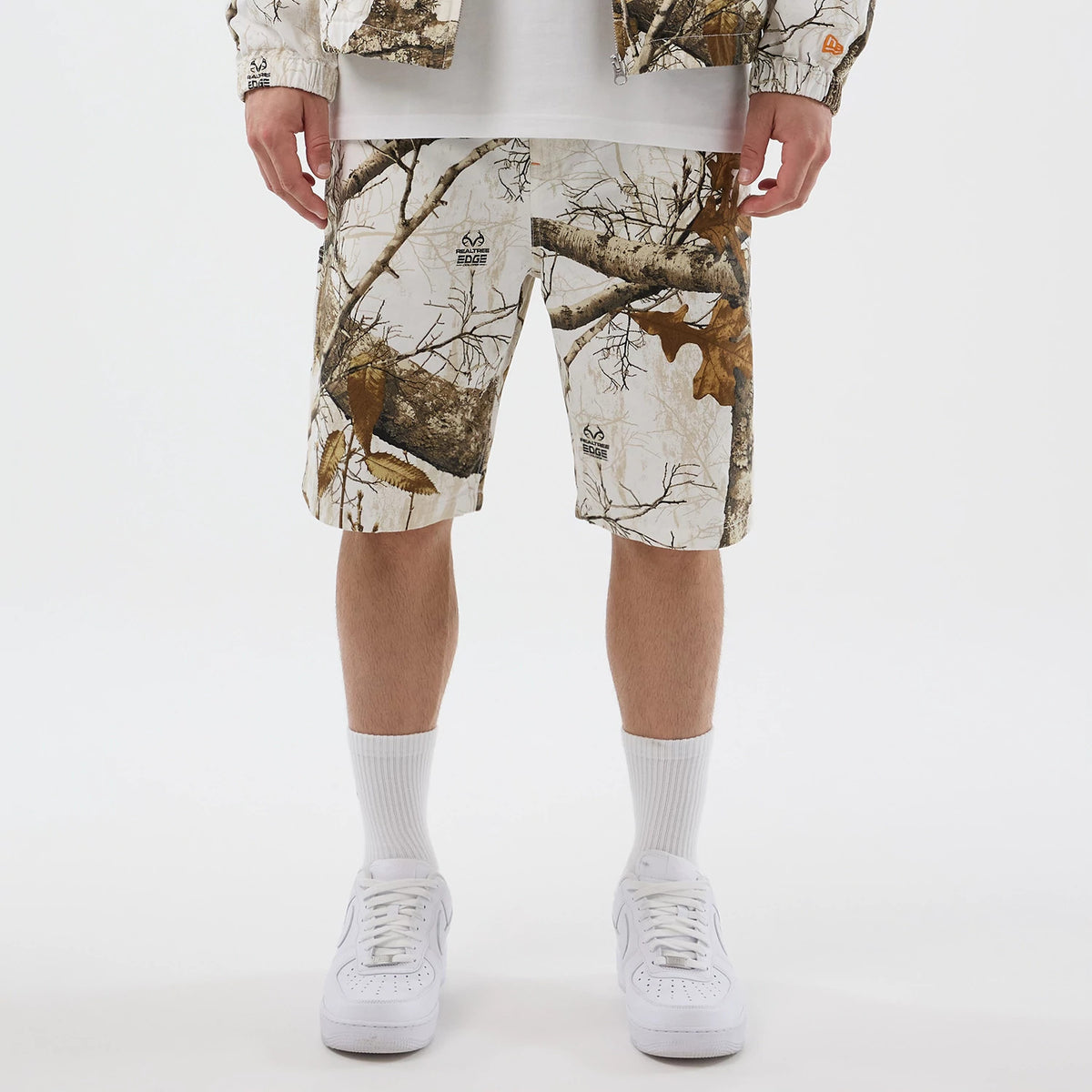 New Era Earthy Skin Real Tree Snow Oversized Shorts, front view showing the camouflage pattern with tree branches and leaves, a debossed New Era logo on the left leg above the knee, worn with white sneakers and socks.