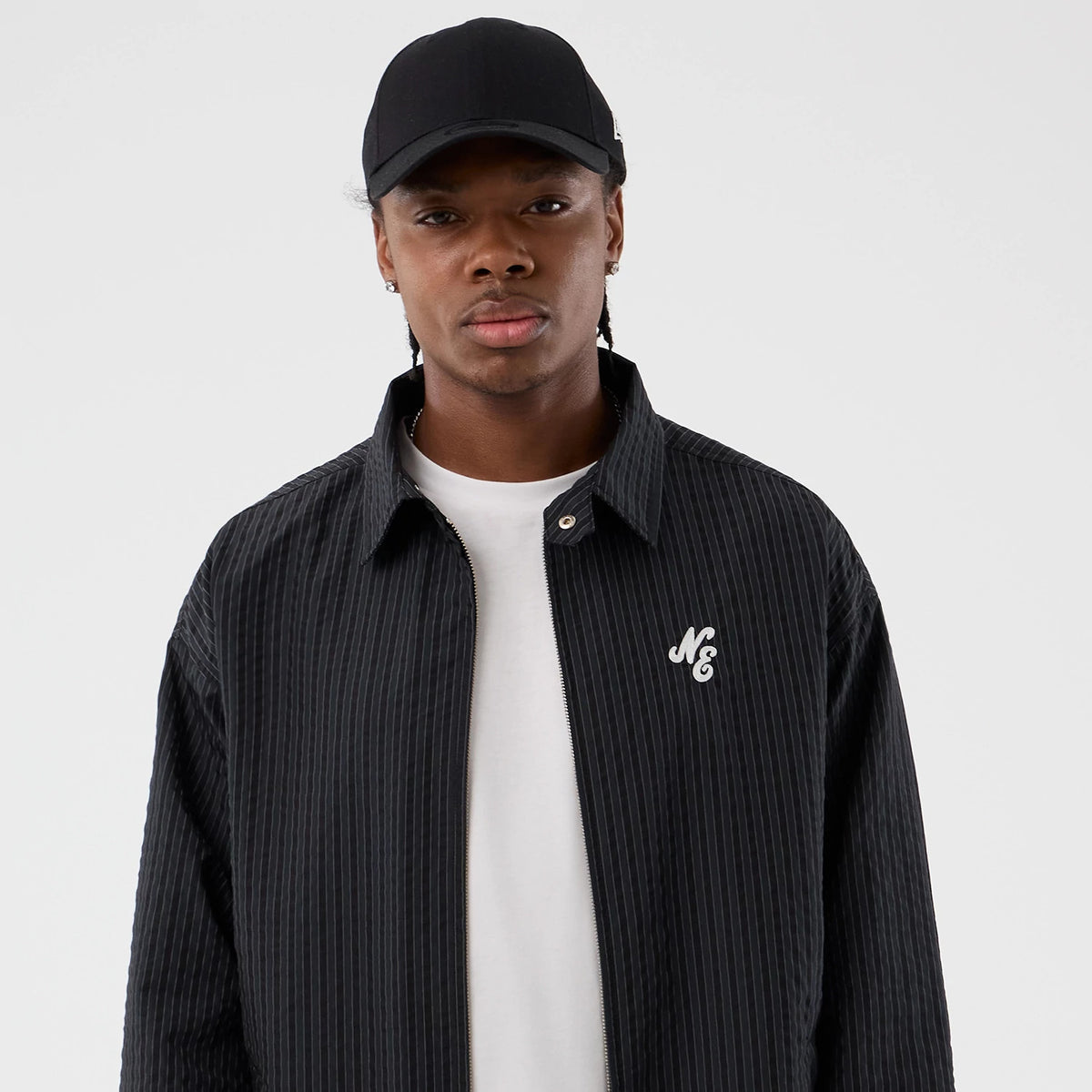 New Era Street Class Navy Jacket, front view showing a pinstriped navy jacket with a raised white "NE" logo on the chest, worn over a white shirt and paired with a plain black cap.