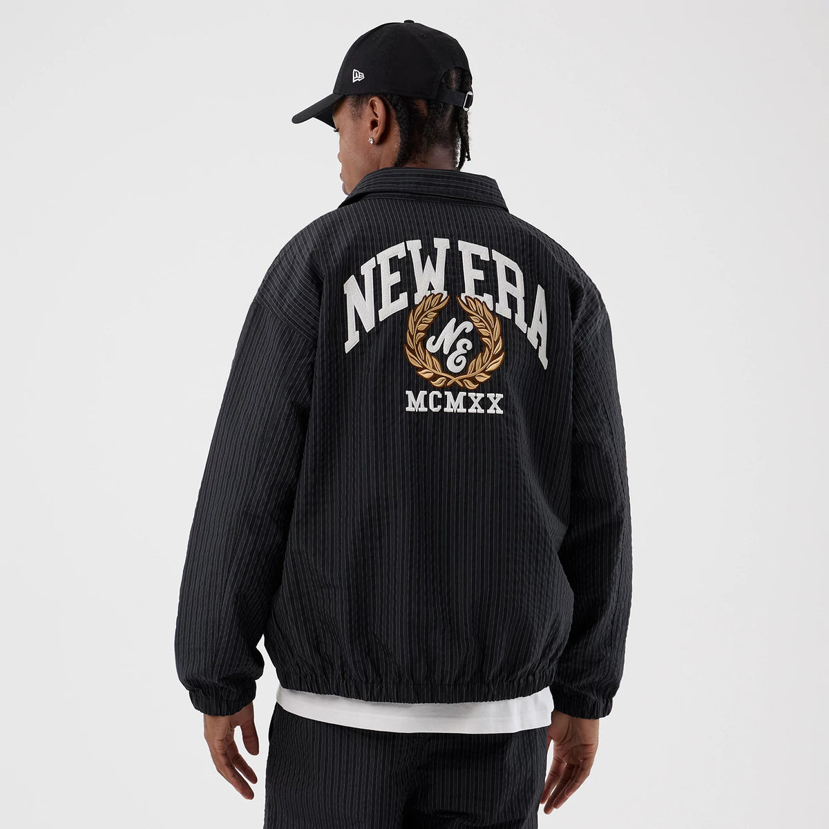 New Era Street Class Navy Jacket, rear view showing a large embroidered New Era logo across the back with decorative laurel wreath and MCMXX detail, worn by a person with a black cap.