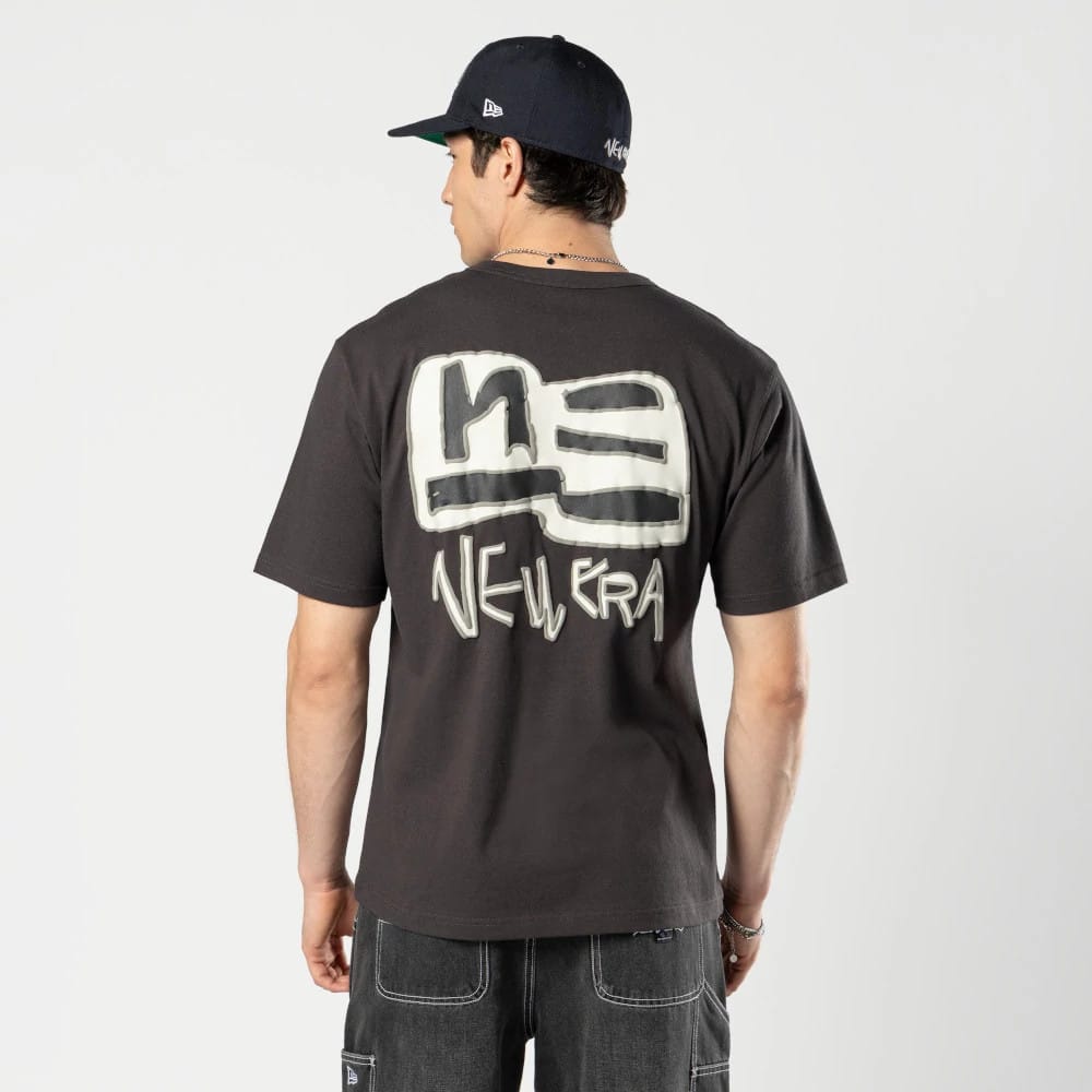 New Era Ochiai Shohei Grey T-Shirt, rear view showing a large stylised logo in white and black on the back, the words "New Era" below, worn by a person also wearing a dark cap with small logos on the side and back.