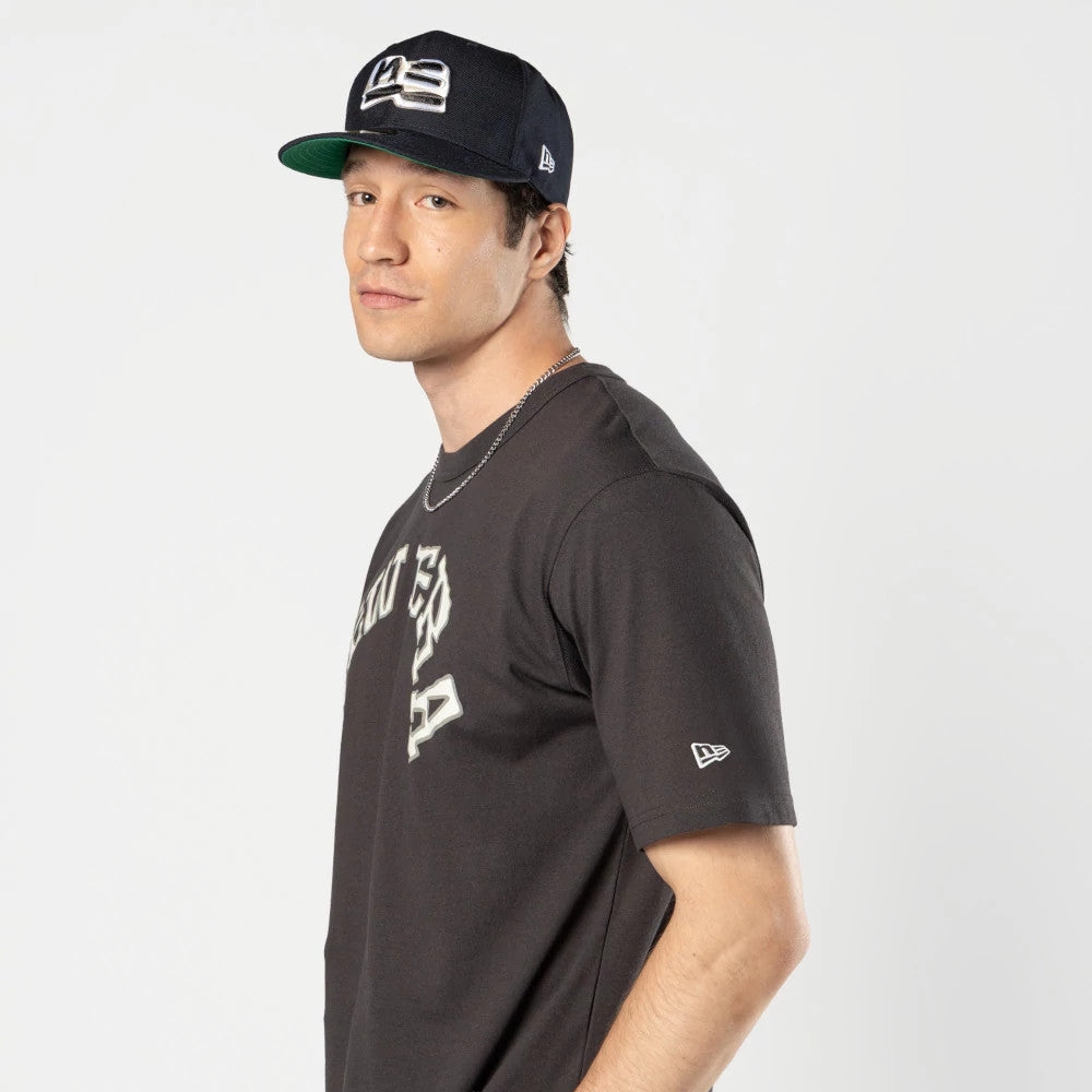 New Era Ochiai Shohei Grey T-Shirt, on-model view showing the grey fabric with white printed letters across the chest, a small raised New Era logo on the sleeve, and a navy cap with another logo worn by the model.