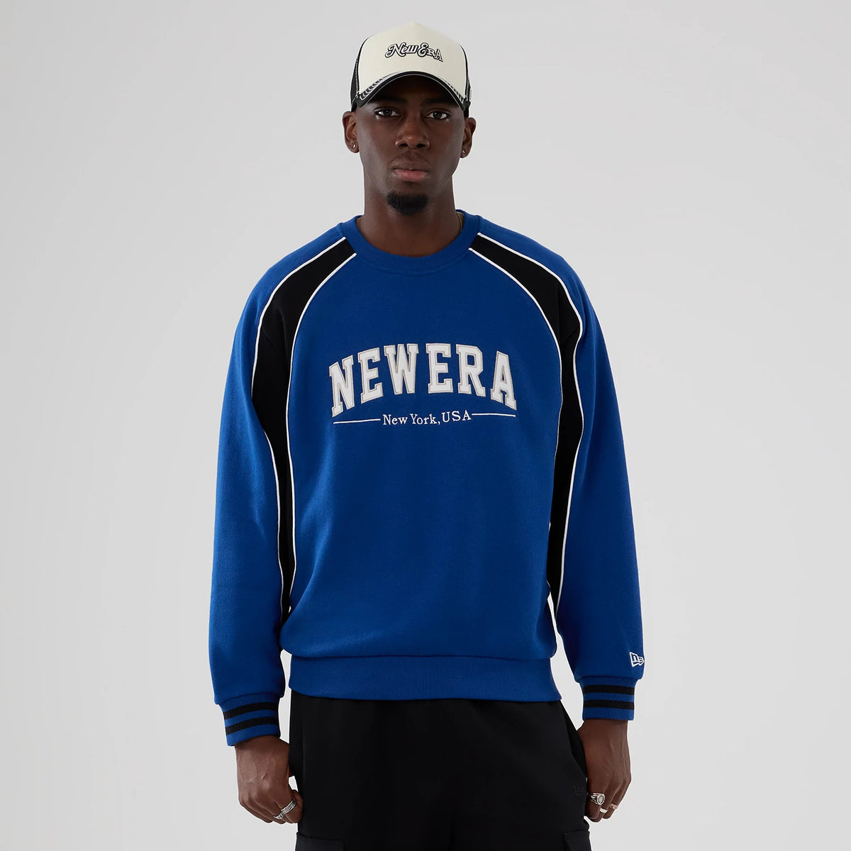 New Era Colour Block Navy Crew Neck Sweater, front view on a model styled with a contrasting cap, featuring large white New Era New York, USA lettering across the chest and black and white panelling on the sleeves.