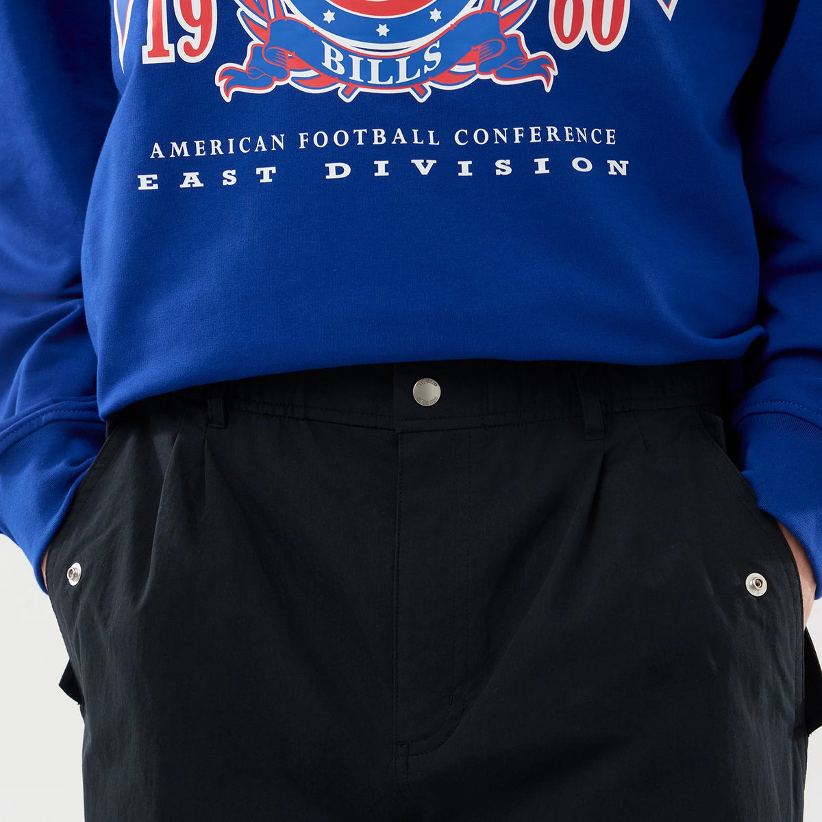 New Era Organic Black Parachute Trousers, close-up on a model wearing a blue sweater, highlighting the trousers' front pleats, silver button closure on the waistband and angled side pockets with matching buttons.