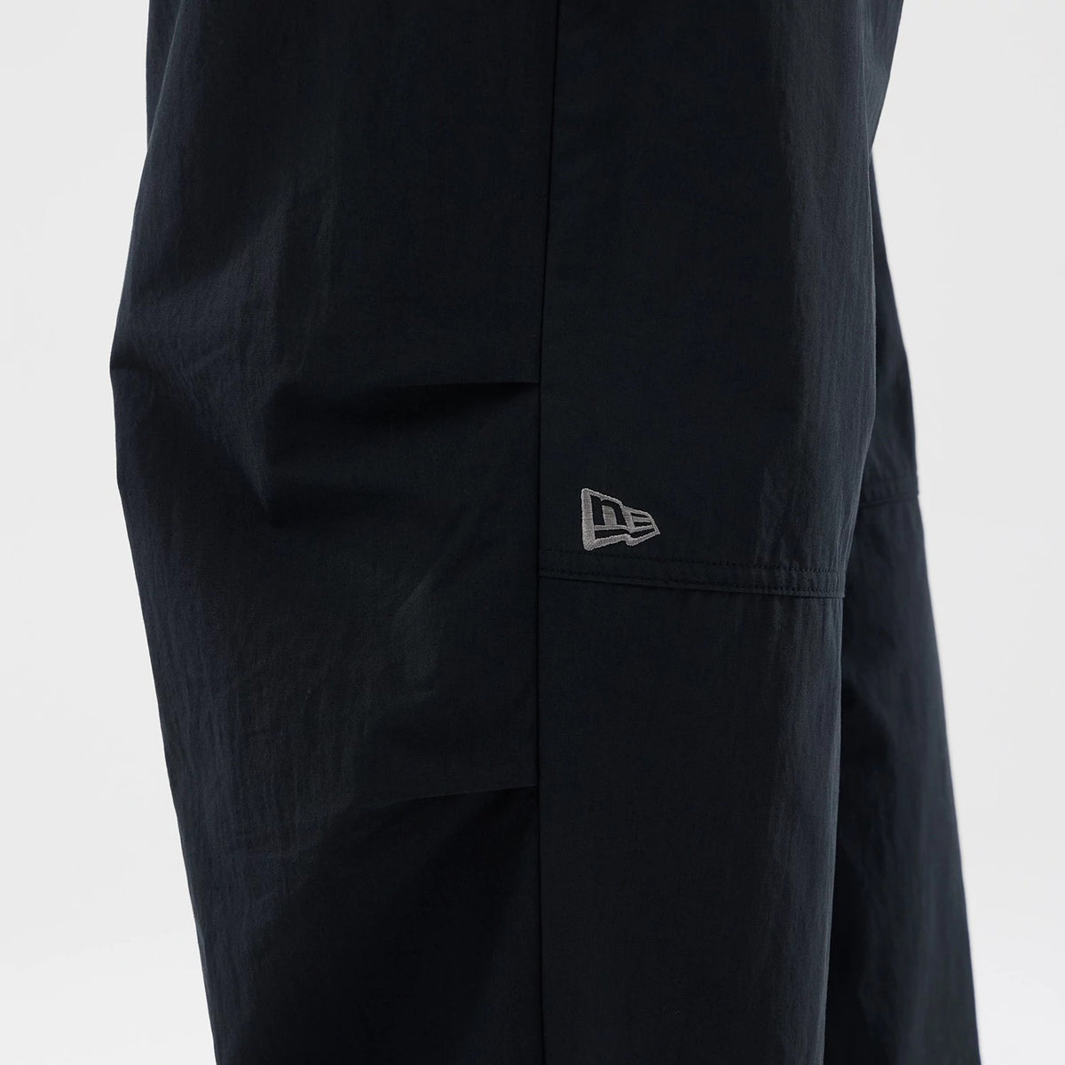 New Era Organic Black Parachute Trousers, side view showing the grey embroidered New Era flag logo positioned below the knee on the relaxed black fabric, highlighting the trousers' loose fit and lightweight design.