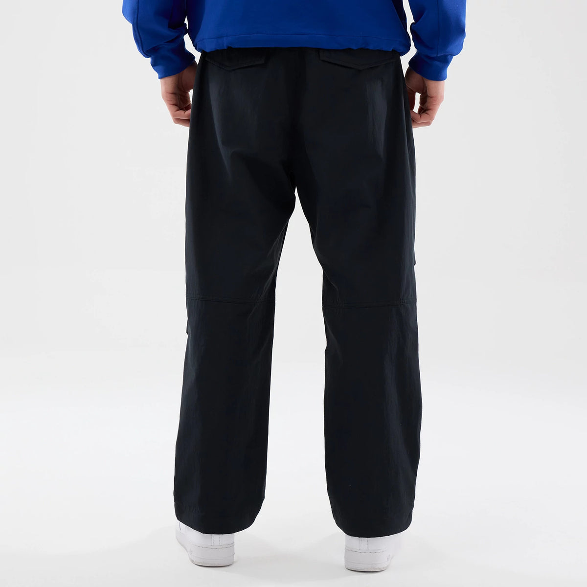 New Era Organic Black Parachute Trousers, back view on a model with a blue top and white shoes, highlighting the relaxed fit and flap pockets on the rear, showcasing the trousers' lightweight design and loose silhouette.