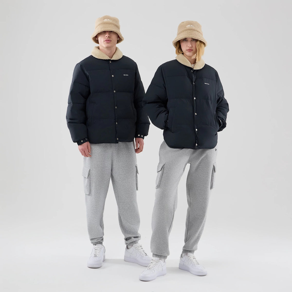 New Era Organic Boa Neck Navy Jacket, front view featuring two models wearing navy jackets with cream collars, button-up fronts and small embroidered New Era logos on the left chest, paired with beige bucket hats and grey trousers.