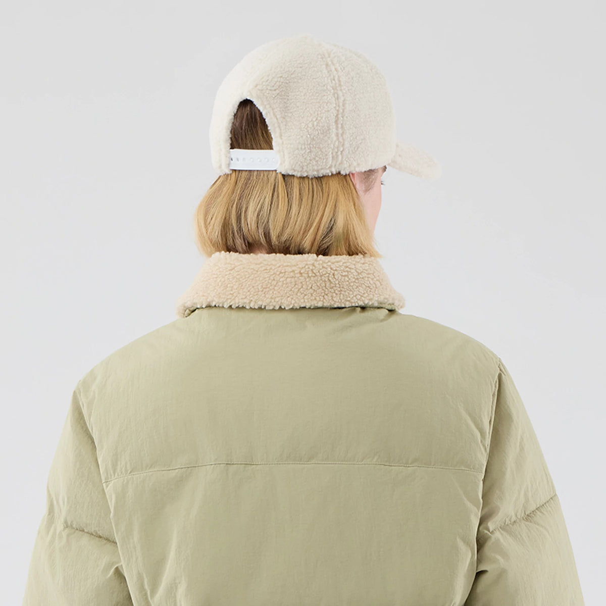 New Era Organic Boa Neck Khaki Jacket, rear view showing the khaki back with a fluffy beige collar, paired with an off-white cap featuring an adjustable strap and a debossed New Era logo on the cap's side.