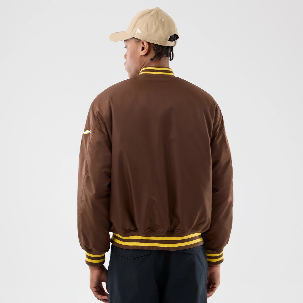 San Diego Padres MLB Authentic Dark Brown Stadium Jacket, rear view showing the dark brown jacket with yellow and brown striped ribbed collar and cuffs, worn by a model in a beige cap with an embossed New Era logo on the side.