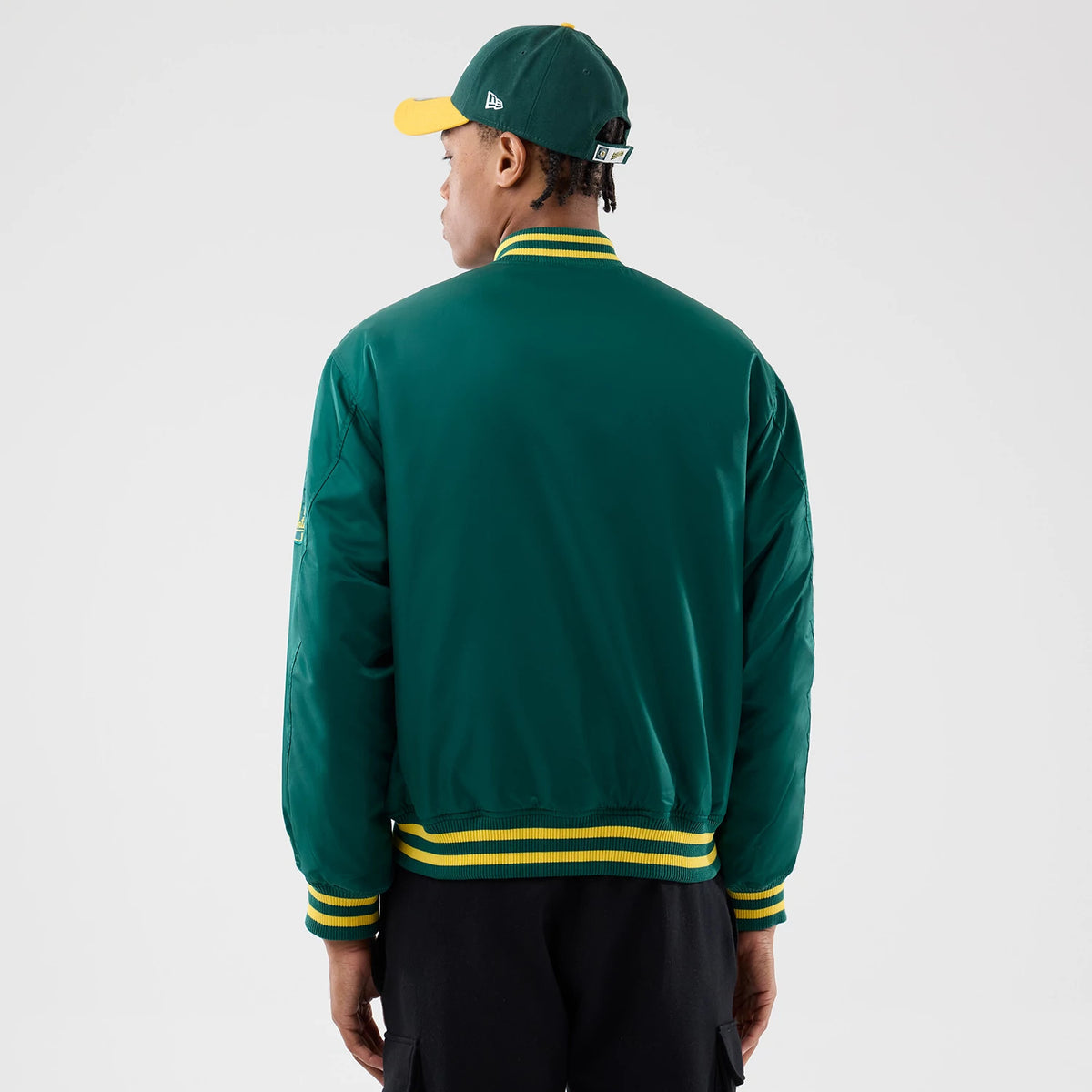 Athletics MLB Authentic Green Stadium Jacket, rear view showing the dark green back with yellow and green striped collar and cuffs, worn by a person in a green cap with a yellow visor, white debossed logo on cap side.