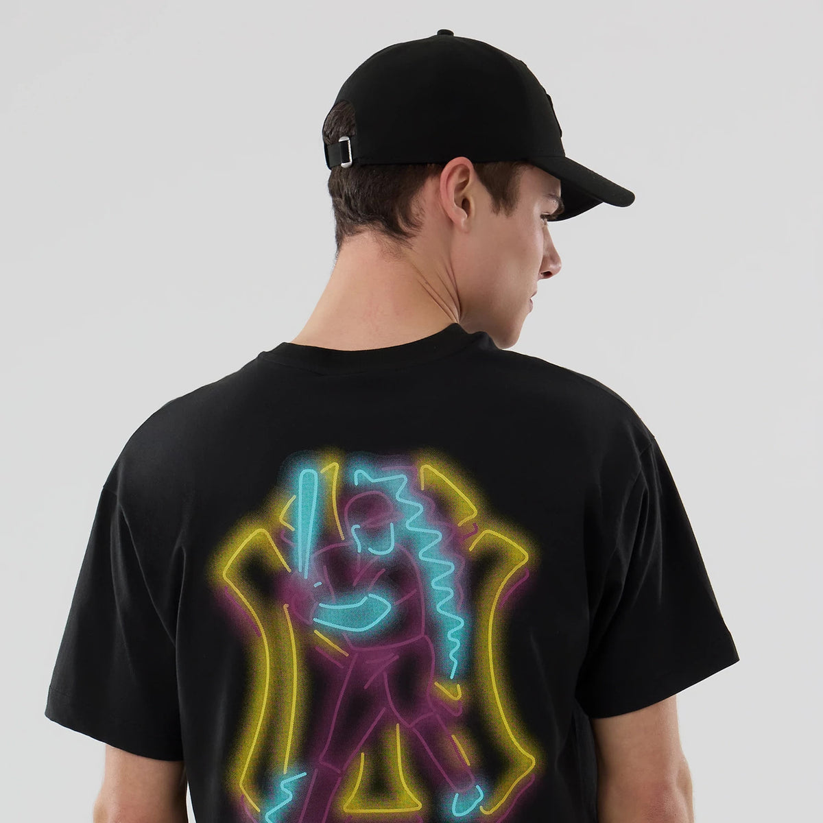 New York Yankees MLB Neon Light Black T-Shirt, on-model view showing the back design featuring a neon-style baseball player graphic in blue, yellow, and pink on a black shirt, worn with a black cap featuring an embossed logo.