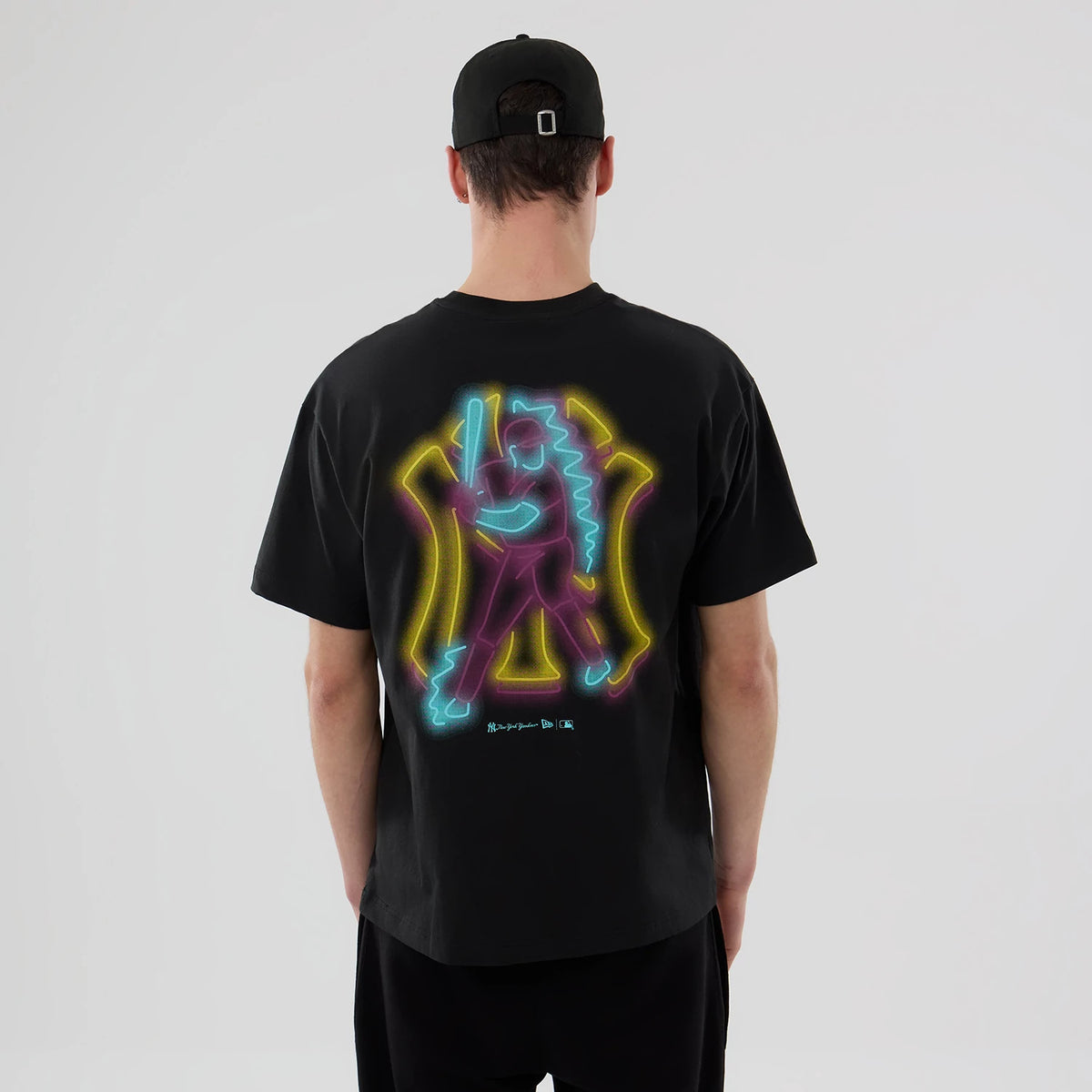 New York Yankees MLB Neon Light Black T-Shirt, back view showing a vibrant neon-style graphic of a baseball player in pink, blue, and yellow on the centre of the black fabric, worn by a person paired with a black cap.