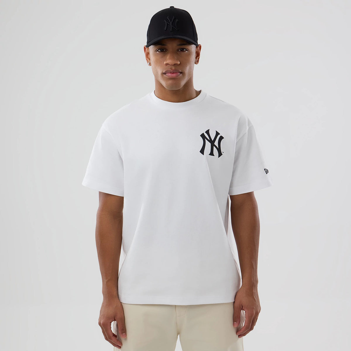 New York Yankees MLB World White T-Shirt, front view showing the white T-shirt with a black New York Yankees logo on the left chest, paired with a black cap featuring a debossed New York Yankees logo on the front.