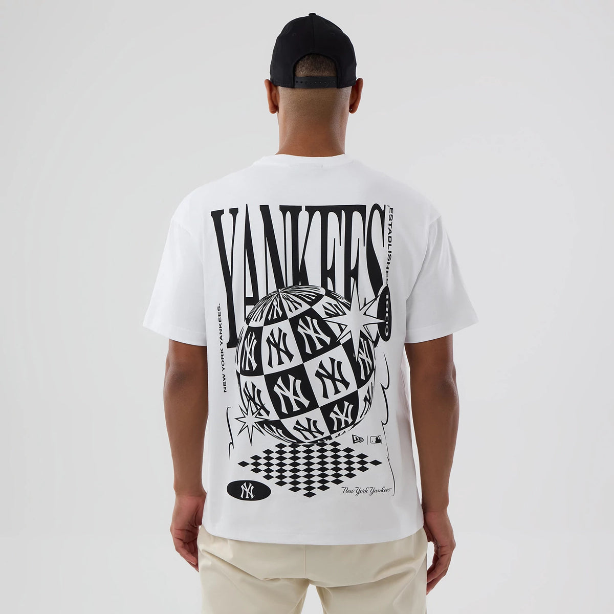 New York Yankees MLB World White T-Shirt, rear view showing a large black graphic design featuring a checkered globe with Yankees logos, bold "YANKEES" text across the back, and small logos at the bottom, worn by a person facing away.