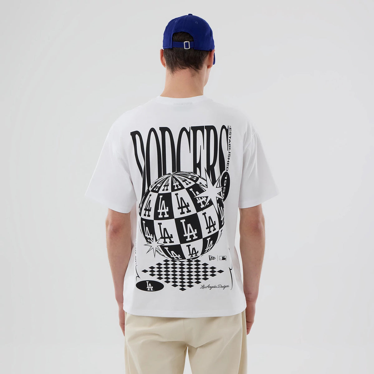 LA Dodgers MLB White T-Shirt, rear view showing large black and white graphic with repeated LA and Dodgers text, a globe design, and additional branding near the hem, worn by a person with a blue cap.