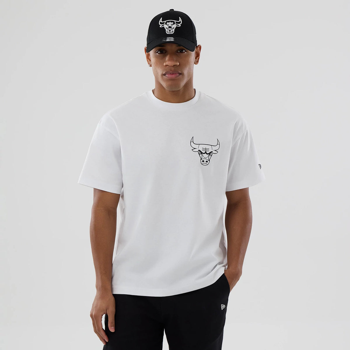 Chicago Bulls NBA World White T-Shirt, on-model view showing a white tee with a black Chicago Bulls logo printed on the left chest, a small embroidered New Era logo on the left sleeve, and a matching logo on a black cap worn by the model.