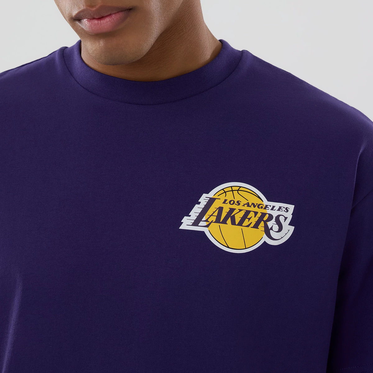 LA Lakers NBA logo Purple T-Shirt, front view showing the purple fabric with the raised Los Angeles Lakers logo in yellow and white on the left chest, featuring a basketball design integrated into the text.