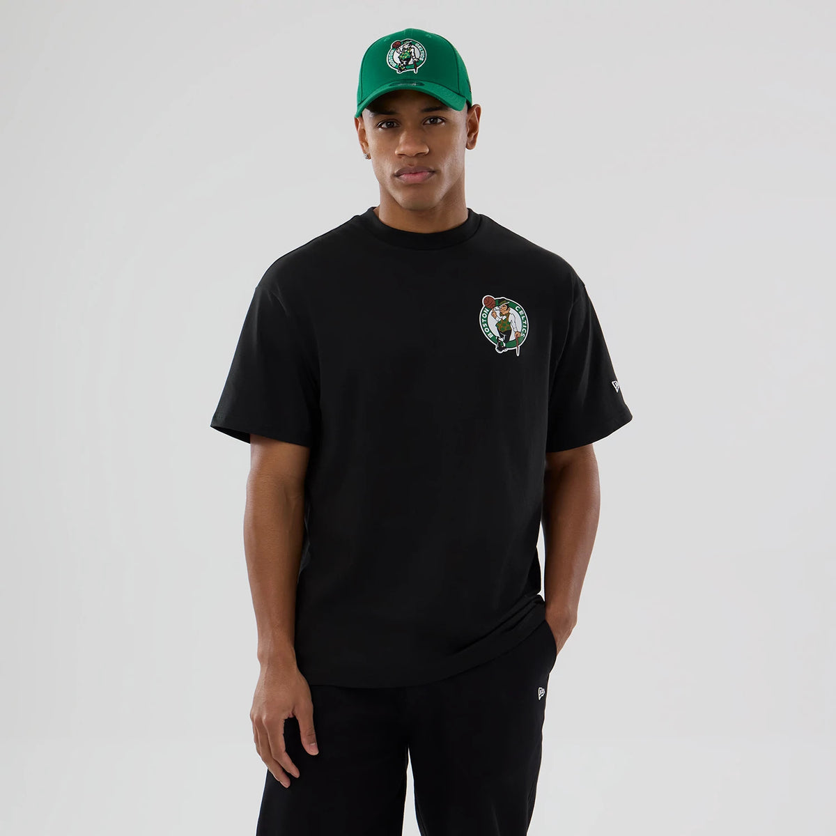 Boston Celtics NBA logo Black T-Shirt, on-model view showing the black T-shirt with a Boston Celtics logo on the left chest, a small debossed New Era logo on the left sleeve, paired with a green cap featuring the same Celtics logo.
